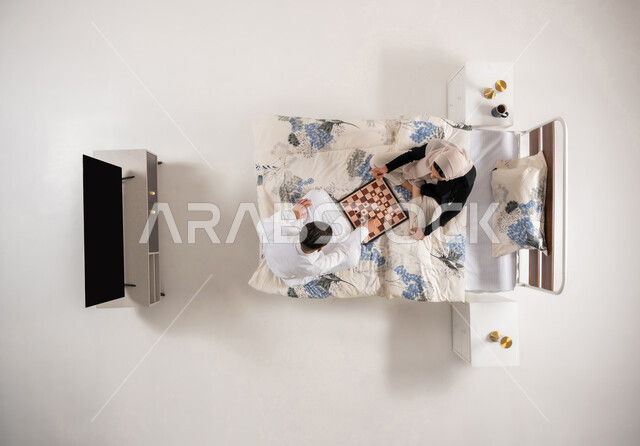 مفهوم التركيز و التحدي, زوجان عمانيان يقضيان وقت ممتع بتعابير التخطيط والتفكير، بورتريه عامودي علوي لإمرأه وشاب عربيان سعوديان خليجيان يجلسان على سرير مريح يلعبان لعبة الشطرنج، ألعاب رقمية استراتيجية، خلفية بيضاء