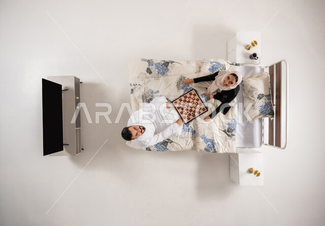 النظر الى الاعلى بإيماءات السرور، مفهوم التركيز و التحدي, زوجان عمانيان يقضيان وقت ممتع بالألعاب الرقمية الاستراتيجية، بورتريه عامودي علوي لإمرأه وشاب عربيان سعوديان خليجيان يجلسان على سرير مريح يلعبان لعبة الشطرنج، ، خلفية بيضاء