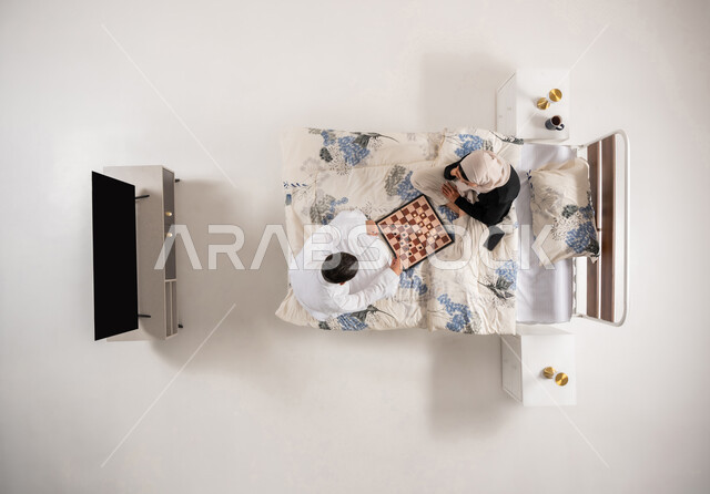 مفهوم التركيز و التحدي, زوجان عمانيان يقضيان وقت ممتع بتعابير التخطيط والتفكير، بورتريه عامودي علوي لإمرأه وشاب عربيان سعوديان خليجيان يجلسان على سرير مريح يلعبان لعبة الشطرنج، ألعاب رقمية استراتيجية، خلفية بيضاء