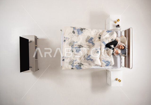 Browsing social media applications, online marketing, vertical top portrait of a veiled Saudi Arabian Gulf woman lying on the bed holding a mobile phone in her hand, an Omani woman chatting with friends with gestures of joy and happiness, white background