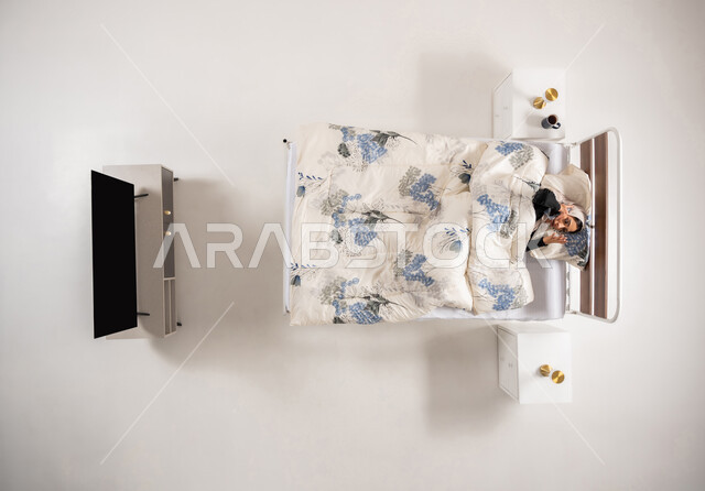 استخدام التقنيات والأجهزة الحديثة، ، بورتريه عامودي علوي لإمرأة عربية خليجية سعودية محجبه مستلقية على السرير تمسك بيدها الهاتف المحمول، سيدة عمانية تقوم بإجراء مكالمة صوتية بتعابير الدهشة والاستغراب، خلفية بيضاء