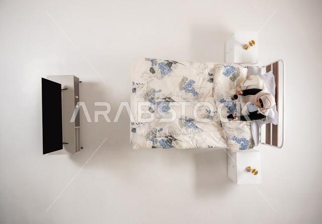 احتساء المشروبات الساخنه، تصفح مواقع التواصل الاجتماعي، سيدة عمانية تستخدم الأجهزة الحديثة، بورتريه عامودي علوي لإمرأة عربية خليجية سعودية مبتسمة ترتدي الحجاب تجلس على سريرها في غرفه النوم تمسك بيدها الهاتف المحمول، الدردشة مع الأهل و الأصدقاء، خلفيه بيضاء