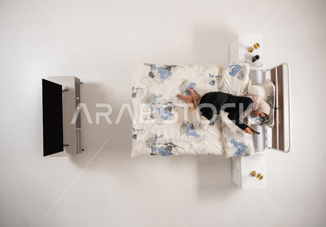 Browsing social media applications, online marketing, vertical top portrait of a veiled Saudi Arabian Gulf woman lying on the bed holding a mobile phone in her hand, an Omani woman chatting with friends with gestures of joy and happiness, white background