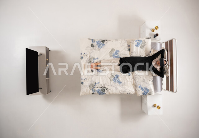 محاكاة العالم الخيالي، امرأة عمانية تستخدم نظارات الواقع الافتراضي بتعابير الاندماج والتركيز، التحول الرقمي والتقدم التقني، بورتريه عامودي علوي لسيده عربية خليجية سعودية محجبه مستلقية على سريرها تضع يديها خلف رأسها تنظر الى الاعلى عبر نظارات الفي ار، خلفيه بيضاء