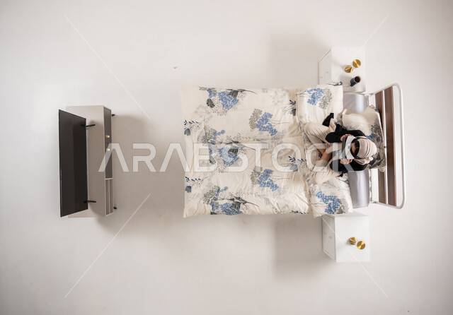 التحول الرقمي والتقدم التقني، محاكاة العالم الخيالي، امرأة عمانية تستخدم نظارات الواقع الافتراضي بتعابير الاندماج والتركيز، بورتريه عامودي علوي لسيده عربية خليجية سعودية محجبه تجلس على سريرها تنظر الى التلفاز من خلال نظارات الفي ار، خلفيه بيضاء