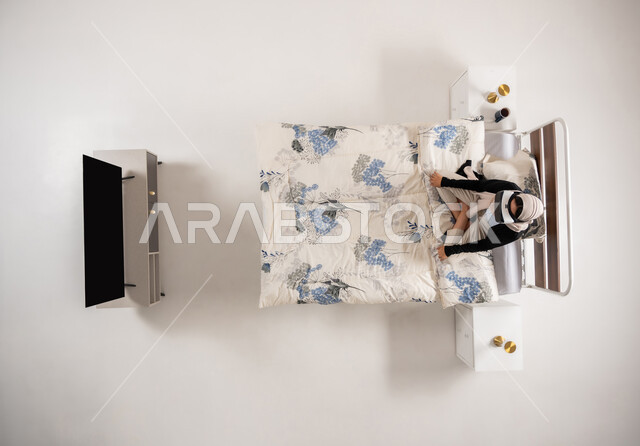 محاكاة العالم الخيالي، امرأة عمانية تستخدم نظارات الواقع الافتراضي بتعابير الاندماج والتركيز، بورتريه عامودي علوي لسيده عربية خليجية سعودية محجبه تجلس على سريرها تنظر الى التلفاز من خلال نظارات الفي ار، التحول الرقمي والتقدم التقني، خلفيه بيضاء