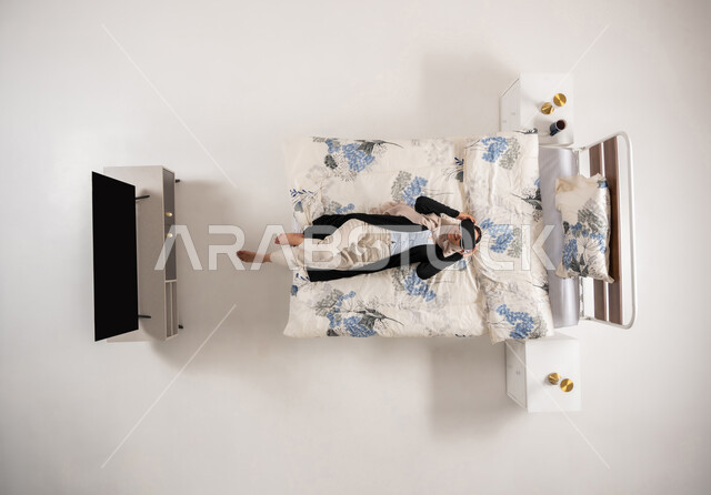 امرأة عمانية تستخدم نظارات الواقع الافتراضي بتعابير الاندماج والتركيز، التحول الرقمي والتقدم التقني، بورتريه عامودي علوي لسيده عربية خليجية سعودية محجبه مستلقية على سريرها تنظر الى الاعلى من خلال نظارات الفي ار، محاكاة العالم الخيالي، خلفيه بيضاء