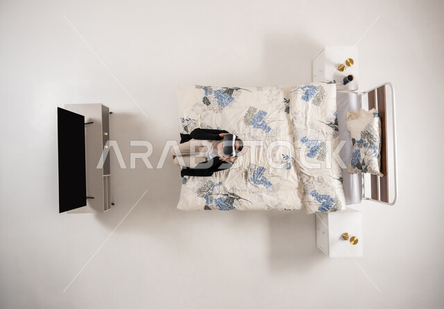 امرأة عمانية تستخدم نظارات الواقع الافتراضي بتعابير الاندماج والتركيز، محاكاة العالم الخيالي، بورتريه عامودي علوي لسيده عربية خليجية سعودية محجبه تجلس على سريرها تنظر الى الاعلى من خلال نظارات الفي ار، التحول الرقمي والتقدم التقني، خلفيه بيضاء