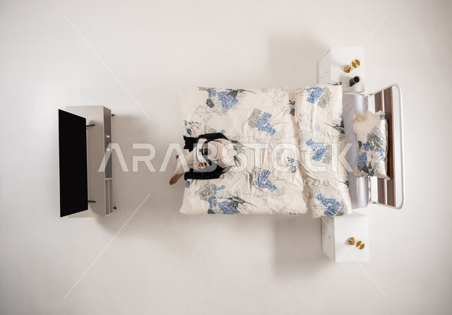 محاكاة العالم الخيالي، امرأة عمانية تستخدم نظارات الواقع الافتراضي بتعابير الاندماج والتركيز، بورتريه عامودي علوي لسيده عربية خليجية سعودية محجبه تجلس على سريرها تنظر الى التلفاز من خلال نظارات الفي ار، التحول الرقمي والتقدم التقني، خلفيه بيضاء