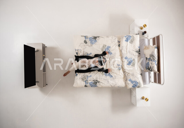 امرأة عمانية تستخدم نظارات الواقع الافتراضي بتعابير الاندماج والتركيز، محاكاة العالم الخيالي، بورتريه عامودي علوي لسيده عربية خليجية سعودية محجبه تجلس على سريرها تنظر الى الاعلى من خلال نظارات الفي ار، التحول الرقمي والتقدم التقني، خلفيه بيضاء