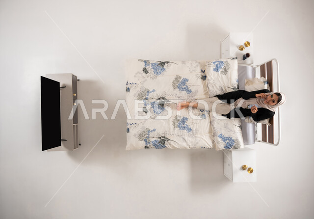 تصفح تطبيقات التواصل الاجتماعي، استخدام التقنيات والأجهزة الحديثة، بورتريه عامودي علوي لإمرأة عربية خليجية سعودية محجبه تجلس على السرير تمسك بيدها الهاتف المحمول تقوم بإجراء مكالمة صوتية، سيدة عمانية تنظر الى الكاميرا بإيماءات السرور، خلفية بيضاء