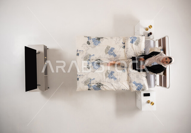 إمرأة عمانية تقوم بإنجاز مهام العمل بإيماءات الاندماج والتركيز، استخدام الأجهزة التكنولوجية الحديثة، بورتريه عامودي علوي لإمرأة عربية خليجية سعودية مبتسمة ترتدي الحجاب تجلس على سريرها في غرفه النوم تنظر الى الكاميرا تعمل على جهاز الحاسب المحمول، خلفيه بيضاء