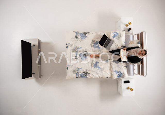 النظر للاعلى بايماءات السرور, الإهتمام بالعلم والثقافه, بورتريه عامودي علوي لإمرأة عربيه سعوديه خليجيه محجبة تجلس على سريرها في غرفة النوم تقوم بتنمية وتعزيز المعلومات العامة, سيدة عمانية تمارس هواية المطالعة والاستكشاف, خلفيه بيضاء