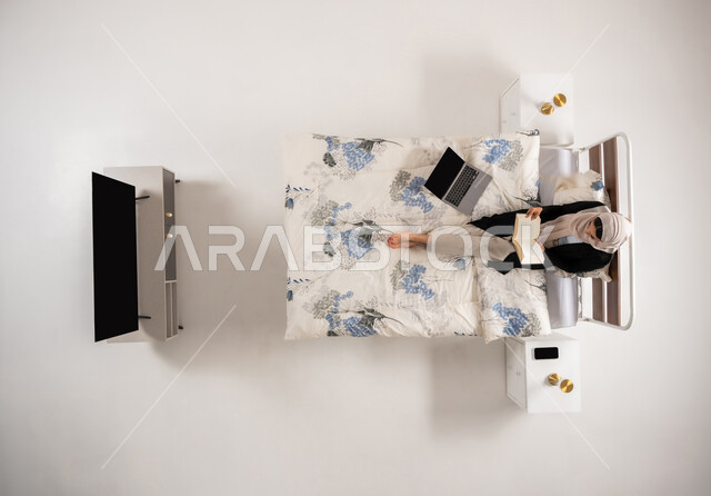 الإهتمام بالعلم والثقافه, بورتريه عامودي علوي لإمرأة عربيه سعوديه خليجيه محجبة تجلس على سريرها في غرفة النوم تقوم بتنمية وتعزيز المعلومات العامة, سيدة عمانية تمارس هواية المطالعة والاستكشاف, خلفيه بيضاء