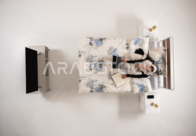 تنمية وتعزيز المعلومات العامة, هواية المطالعة والاستكشاف، بورتريه عامودي علوي لإمرأة عربيه سعوديه خليجيه محجبة تجلس على سريرها في غرفة النوم تقوم بالبحث عن رواية عبر الحاسوب المحمول, سيدة عمانية تنظر للاعلى بتعابير السرور, الإهتمام بالعلم والثقافه, خلفيه بيضاء