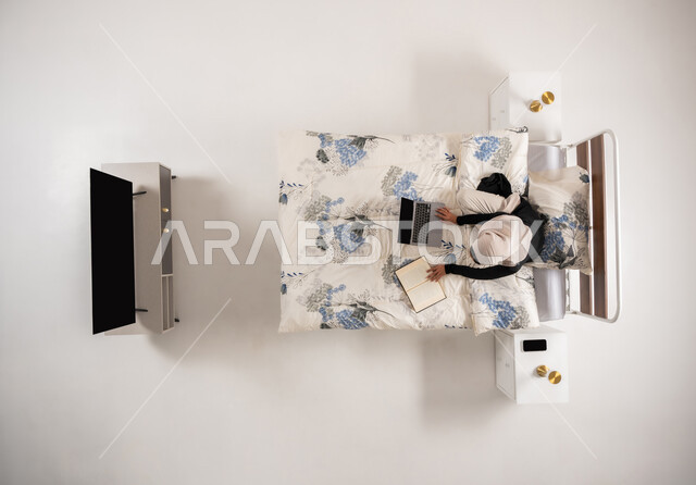 هواية المطالعة والاستكشاف، تنمية وتعزيز المعلومات العامة, بورتريه عامودي علوي لإمرأة عربيه سعوديه خليجيه محجبة تجلس على سريرها في غرفة النوم تقوم بالبحث عن رواية عبر الحاسوب المحمول, سيدة عمانية تقضي وقت ممتع في القراءة, الإهتمام بالعلم والثقافه, خلفيه بيضاء