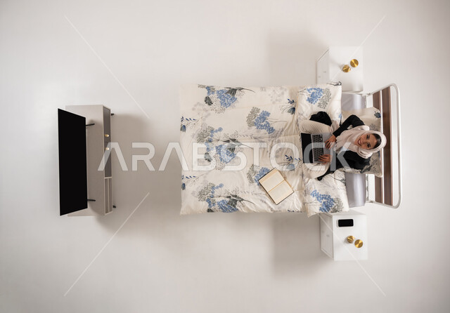 Looking up with expressions of pleasure, using modern advanced technologies, Omani lady sitting on her bed in bedroom searching for online bookstores, vertical top portrait of veiled Saudi Arabian Gulf woman working on laptop, white background