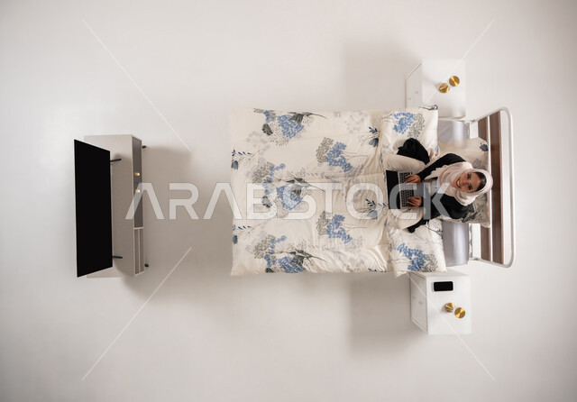 النظر الى الاعلى بتعابير السرور، التواصل مع الاهل والأصدقاء، سيدة عمانية تجلس على سريرها في غرفة النوم تستخدم التقنيات الحديثة المتطورة والتكنولوجيا, بورتريه علوي عامودي لامرأة عربيه خليجيه سعوديه محجبه تقوم بالعمل على جهاز الحاسوب المحمول، خلفية بيضاء