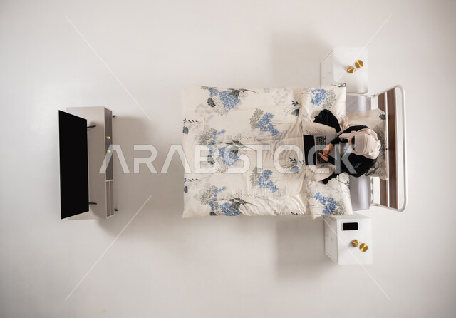 استخدام التقنيات الحديثة المتطورة والتكنولوجيا، التواصل مع الاهل والأصدقاء، سيدة عمانية تجلس على سريرها في غرفة النوم تقوم بإدارة وتنظيم الاعمال عن بعد، بورتريه علوي عامودي لامرأة عربيه خليجيه سعوديه محجبه تقوم بالعمل على جهاز الحاسوب المحمول، خلفية بيضاء