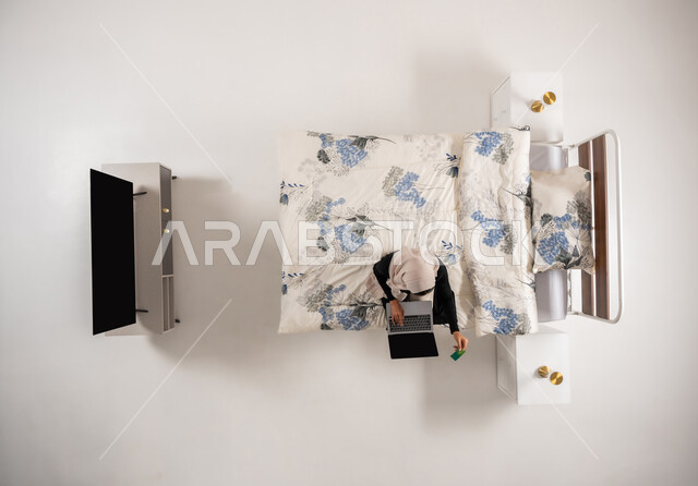 استخدام بطاقة الإئتمان في التسوق الالكتروني، الشراء والدفع عن بعد عبر الاونلاين, بورتريه علوي عامودي لسيدة عربية سعودية خليجية محجبة تجلس على سريرها في غرفة النوم تمسك البطاقة البنكية تقوم بتصفح تطبيقات المصارف والبنوك عبر جهاز الحاسوب المحمول، خلفية بيضاء