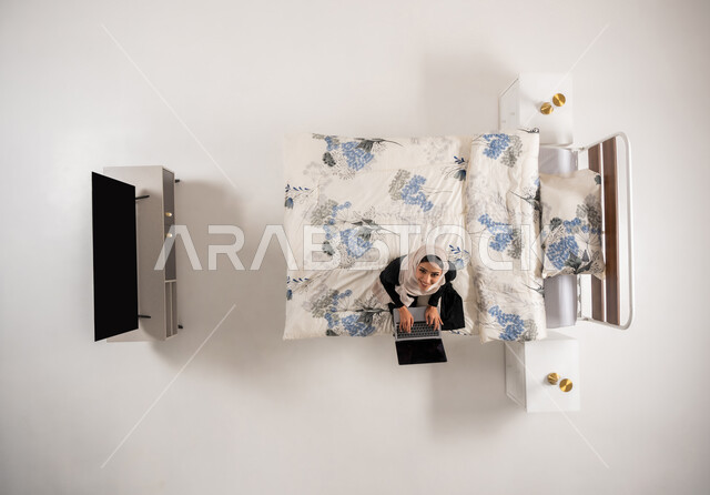 Using modern advanced techniques and technology, communicating with family and friends, Omani woman sitting on her bed in the bedroom looking up with expressions of pleasure, vertical top portrait of a veiled Saudi Arabian Gulf woman working on a laptop, white background
