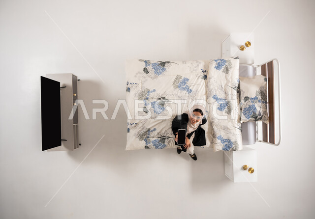 Using modern technical devices, vertical overhead portrait of a veiled Saudi Arabian Gulf woman wearing a black abaya holding a mobile phone looking at the camera with gestures of pleasure, Omani woman sitting on her bed in the bedroom showing a blank black screen via mobile, white background