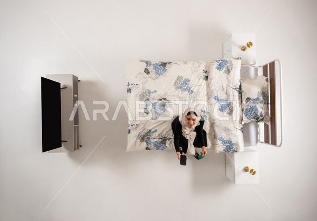 Online payment and purchase, Omani woman holding bank card, using credit card in online shopping, vertical top portrait of veiled Saudi Arabian Gulf woman sitting on her bed in bedroom browsing banking applications via mobile, white background