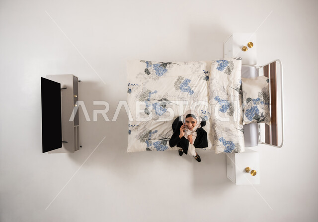 قضاء اوقات ممتعة بتبادل الاخبار والمحادثات عن بعد، بورتريه علوي عامودي لسيده عربية خليجية عمانيه محجبة تقوم باجراء احاديث ودردشه مع الاهل والاصدقاء باستخدام الهاتف المحمول، امرأة سعودية تجلس على سريريها وتنظر الى الاعلى بتعابير السرور، خلفيه بيضاء