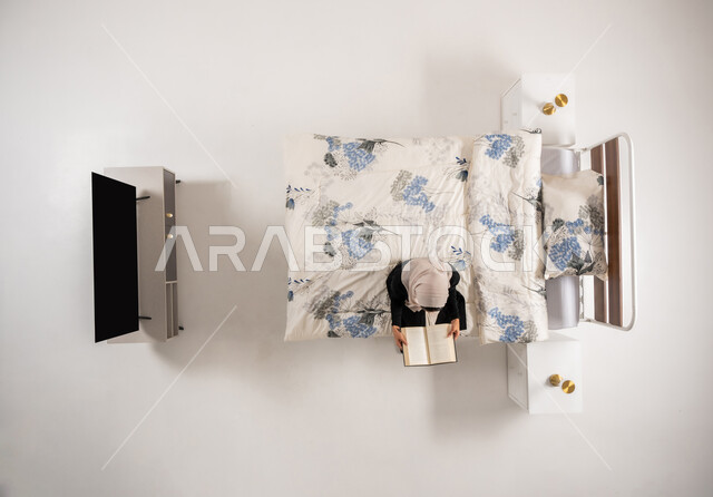 تنمية وتعزيز المعلومات العامة, هواية المطالعة والاستكشاف، سيدة عمانية تقضي وقت ممتع في القراءة, بورتريه عامودي علوي لإمرأة عربيه سعوديه خليجيه محجبة ترتدي الكاجوال تجلس على سريرها في غرفة النوم تمسك كتاب, الإهتمام بالعلم والثقافه, خلفيه بيضاء