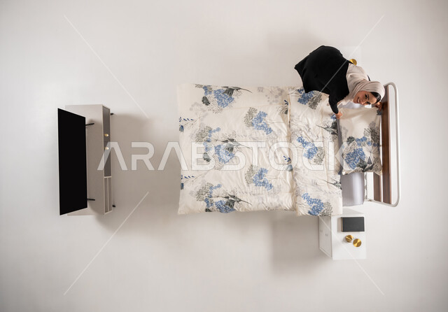 النظر الى الكاميرا بإيماءات النشاط والسرور، مفهوم التدبير المنزلي و القيام بالأعمال الروتينية، امرأة عمانية تهتم بتنظيم المنزل، بورتريه عامودي علوي لسيدة عربية سعودية خليجية محجبة تقوم بترتيب سريرها في غرفة النوم، خلفيه بيضاء