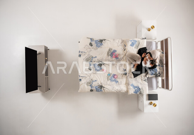 سيدة عمانية تعانق طفلتها الصغيرة بإيماءات الحب والحنان، تعزيز الروابط الأسرية، الرعاية والاهتمام بالأبناء, بورتريه عامودي علوي لأم عربيه خليجيه سعوديه محجبة تحمل ابنتها حديثه الولاده تجلس على سريرها, خلفيه بيضاء