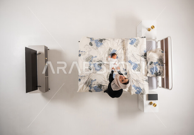 سيدة عمانية تنظر الى الكاميرا بتعابير السرور تداعب طفلتها الصغيرة، الرعاية والاهتمام بالأبناء, النظر إلى الاطفال بإيماءات الحب والحنان، بورتريه عامودي علوي لأم عربيه خليجيه سعوديه محجبة تلعب مع ابنتها حديثه الولاده على سريرها, خلفيه بيضاء