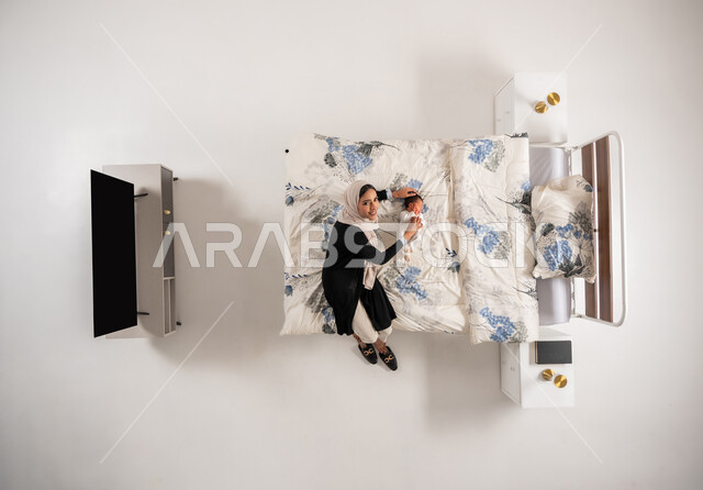 سيدة عمانية تنظر الى الكاميرا بتعابير السرور تداعب طفلتها الصغيرة، الرعاية والاهتمام بالأبناء, النظر إلى الاطفال بإيماءات الحب والحنان، بورتريه عامودي علوي لأم عربيه خليجيه سعوديه محجبة تلعب مع ابنتها حديثه الولاده على سريرها, خلفيه بيضاء