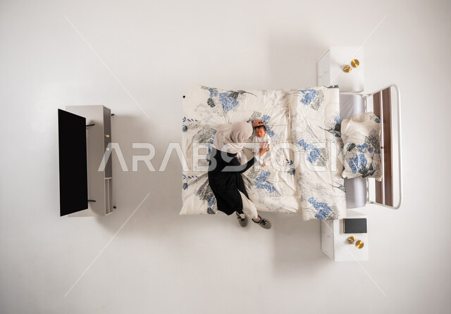 Looking at children with gestures of love and tenderness, Omani woman caressing her little girl, vertical top portrait of a veiled Saudi Arabian Gulf mother playing with her newborn daughter on her bed, baby wearing a soft cotton rompers, care and attention for children, white background