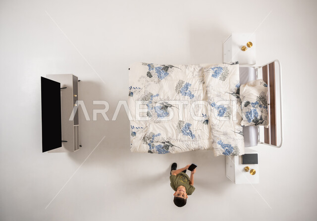 صبي عماني يمسك بيده جهاز الهاتف المحمول, استعمال الأجهزة التكنولوجية الحديثة والمتطورة, تصفح مواقع التواصل الاجتماعي، بورتريه علوي عامودي لولد عربي خليجي سعودي يقف امام السرير في غرفه النوم ينظر الى الكاميرا يقوم باستخدام الجوال, خلفية بيضاء