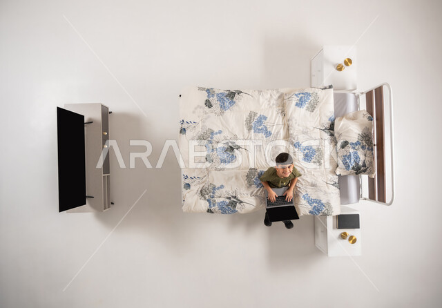 صبي عماني يمسك بيده الحاسب المحمول, استعمال الأجهزة التكنولوجية الحديثة والمتطورة, تصفح مواقع التواصل الاجتماعي، بورتريه علوي عامودي لولد عربي خليجي سعودي يجلس على السرير في غرفه النوم ينظر الى الكاميرا يقوم باستخدام جهاز اللابتوب, خلفية بيضاء