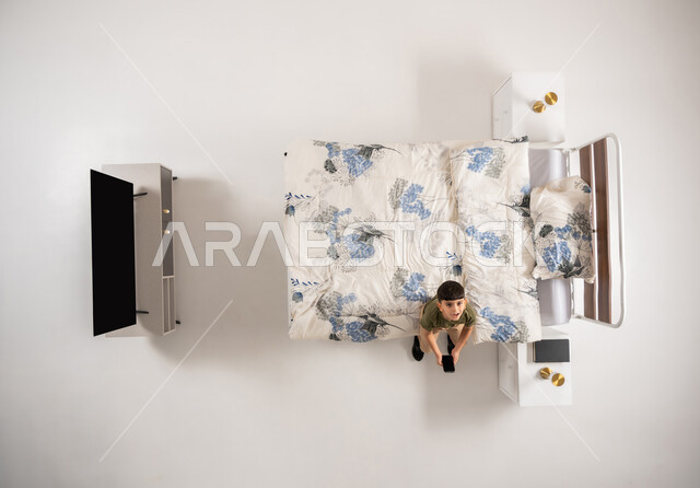 صبي عماني يمسك بيده جهاز الهاتف المحمول, استعمال الأجهزة التكنولوجية الحديثة والمتطورة, بورتريه علوي عامودي لولد عربي خليجي سعودي يجلس على السرير في غرفه النوم ينظر الى الكاميرا بإيماءات السرور, تصفح مواقع التواصل الاجتماعي، خلفية بيضاء