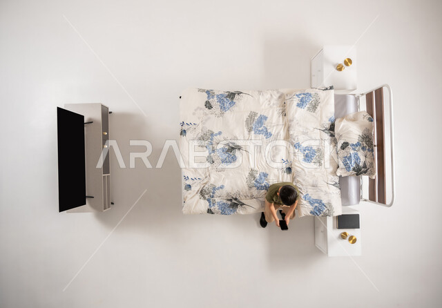 Browsing social media, Omani boy holding mobile phone, using modern and advanced technological devices, vertical overhead portrait of a Saudi Arabian Gulf boy sitting on the bed in his bedroom, white background