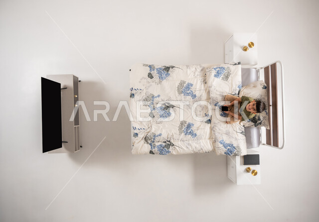 صبي عماني يمسك بيده جهاز الهاتف المحمول, استعمال الأجهزة التكنولوجية الحديثة والمتطورة, بورتريه علوي عامودي لولد عربي خليجي سعودي يجلس على السرير في غرفه النوم ينظر الى الكاميرا بإيماءات السرور, تصفح مواقع التواصل الاجتماعي، خلفية بيضاء