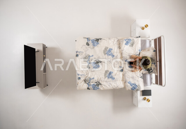 تصفح مواقع التواصل الاجتماعي، صبي عماني يمسك بيده جهاز الهاتف المحمول, استعمال الأجهزة التكنولوجية الحديثة والمتطورة, بورتريه علوي عامودي لولد عربي خليجي سعودي يجلس على السرير في غرفه النوم, خلفية بيضاء