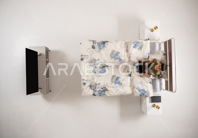 Using modern and advanced technological devices, vertical overhead portrait of a Saudi Arabian Gulf boy sitting on the bed in his bedroom looking at the camera with gestures of pleasure, Omani boy holding a portable tablet in his hand, white background