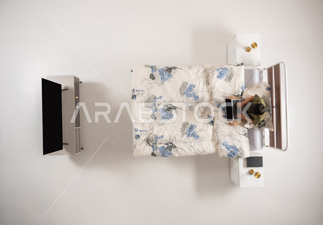 استعمال الأجهزة التكنولوجية الحديثة والمتطورة, بورتريه علوي عامودي لولد عربي خليجي سعودي يجلس على السرير في غرفه النوم, صبي عماني يقوم باستخدام جهاز التابليت المحمول بتعابير وايماءات الاندماج والتركيز, خلفية بيضاء