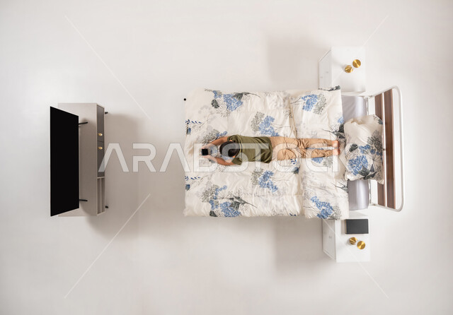 استخدام التقنيات والأجهزة الحديثة، ولد عماني يستخدم جهاز الهاتف المحمول، الاسترخاء على سرير ناعم، بورتريه عامودي علوي لصبي عربي خليجي سعودي مستلق على بطنه على سريره، تصفح تطبيقات و برامج التواصل الاجتماعي، خلفيه بيضاء