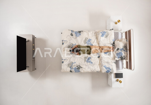 استخدام التقنيات والأجهزة الحديثة، ولد عماني يستخدم جهاز الهاتف المحمول، الاسترخاء على سرير ناعم، بورتريه عامودي علوي لصبي عربي خليجي سعودي مستلق على بطنه على سريره، تصفح تطبيقات و برامج التواصل الاجتماعي، خلفيه بيضاء