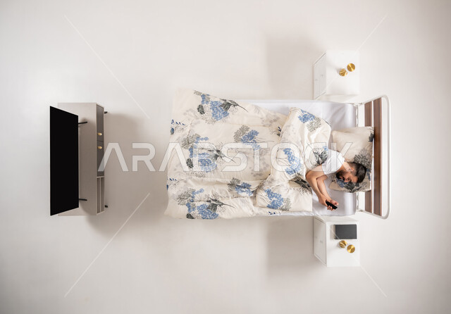 استخدام التقنيات والأجهزة الحديثة، شاب عماني يستخدم جهاز الهاتف المحمول، الاسترخاء والراحة على سرير ناعم وقت القيلولة، بورتريه عامودي علوي لرجل عربي خليجي سعودي مستلق على سريره، الدردشة مع الأهل والاصدقاء عبر برامج التواصل الاجتماعي، خلفيه بيضاء