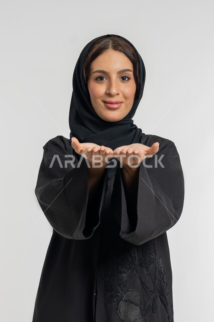 شابة اماراتية تقوم بحمل ورفع عنصر ما على الكفين، النظر إلى الكاميرا بايماءات السعادة والسرور، بورتريه لامرأة عربية خليجية سعودية محجبة ترتدي عباءة سوداء تفتح يديها لعرض شيء ما، خلفية بيضاء