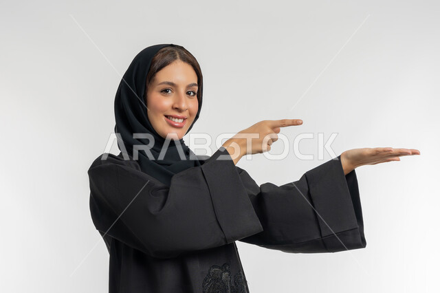 الاشارة الى عنصر هولوجرامي، رفع شيء معين, بورتريه مقرب من الجانب لامرأة عربية خليجية إماراتية محجبة ترتدي العباءة السوداء ترفع كفها للأعلى وتشير اليه, شابة سعودية مبتسمة تقف بتعابير حمل و إمساك عنصر محدد، خلفية بيضاء