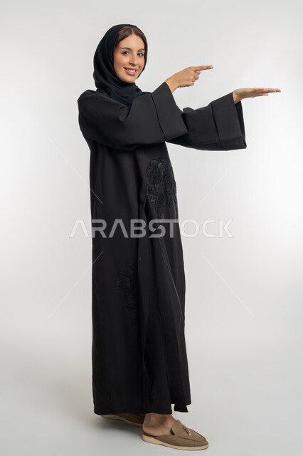 الاشارة الى عنصر هولوجرامي، رفع شيء معين, بورتريه من الجانب لامرأة عربية خليجية إماراتية محجبة ترتدي العباءة السوداء ترفع كفها للأعلى وتشير اليه, شابة سعودية مبتسمة تقف بتعابير حمل و إمساك عنصر محدد, صورة الجسم بالطول الكامل، خلفية بيضاء