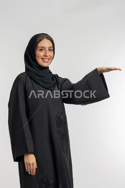 رفع شيء معين, بورتريه من الجانب لامرأة عربية خليجية إماراتية محجبة ترتدي العباءة السوداء ترفع كفها للأعلى, شابة سعودية مبتسمة تقف بتعابير حمل و إمساك عنصر محدد, خلفية بيضاء