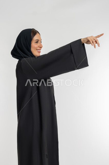 Pointing to an item, explaining and indicating a specific place, side portrait of a veiled Emirati Gulf Arab woman wearing a black abaya pointing up with index finger, young Saudi woman locating something, white background